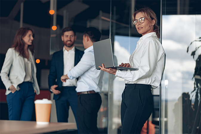 Mature confident businesswoman at office with group of colleagues on background, working on laptop. Teamwork and business concept