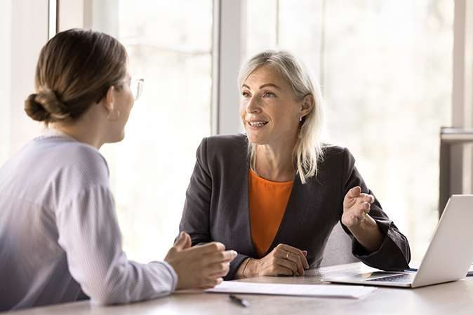 Professional older woman speaking with a younger woman in front of a laptop in an office