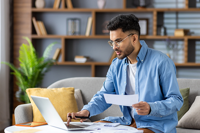 Young man working from home with laptop and documents in modern living room. Casual setting with wooden shelves, plants, and cozy sofa. Focused on work and productivity.