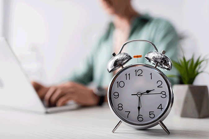 Clock sitting on a desk with the time of two thirty and someone on a laptop behind it