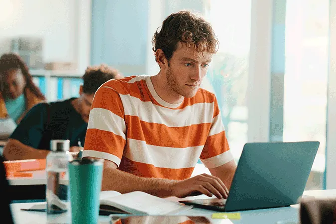 Young man studying on his laptop in a common room with other students