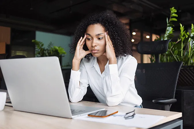 Young woman holding her head in an office sitting in front of a laptop