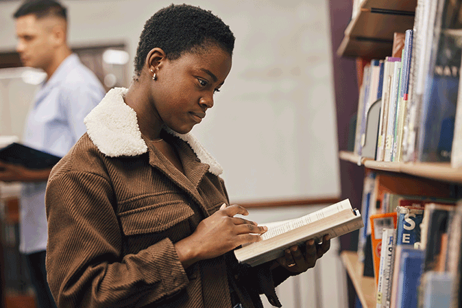 Young woman standing in a library reading a book off a bookshelf