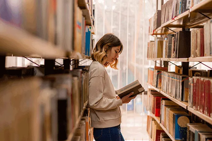 Young woman leaning against a bookcase in a library reading a book
