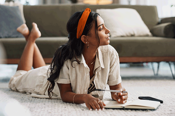Young woman laying on floor in living room thoughtfully writing in notebook