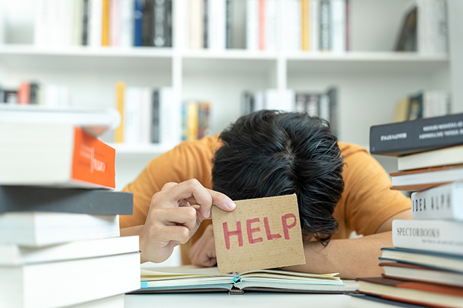 Frustrated students with head down on books holding a help sign