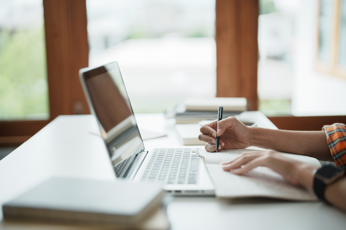 Someone sitting at a desk writing notes in front of a laptop