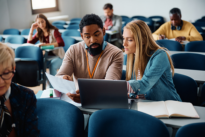 Male professor sitting at a desk in a classroom working with a young woman on an assignment in front of a laptop