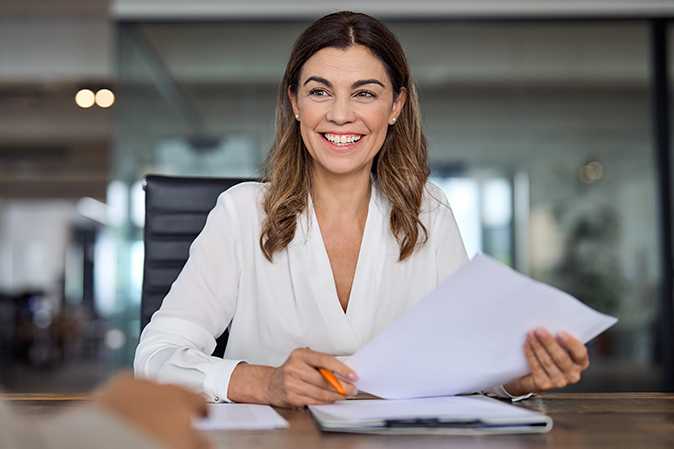 Professional woman smiling while holding documents sitting at a desk in an office