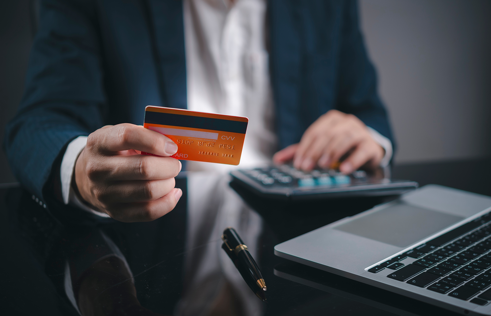 Business man holding a credit card while using a calculator sitting in front of a laptop