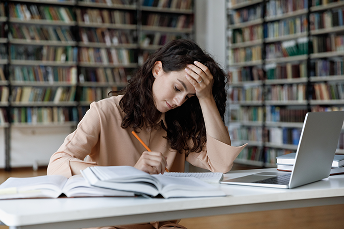 Stressed young woman in a library with books and a laptop writing an essay