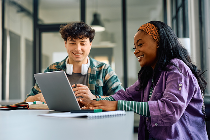 Young woman typing on a laptop with a young man wearing headphones sitting next to her