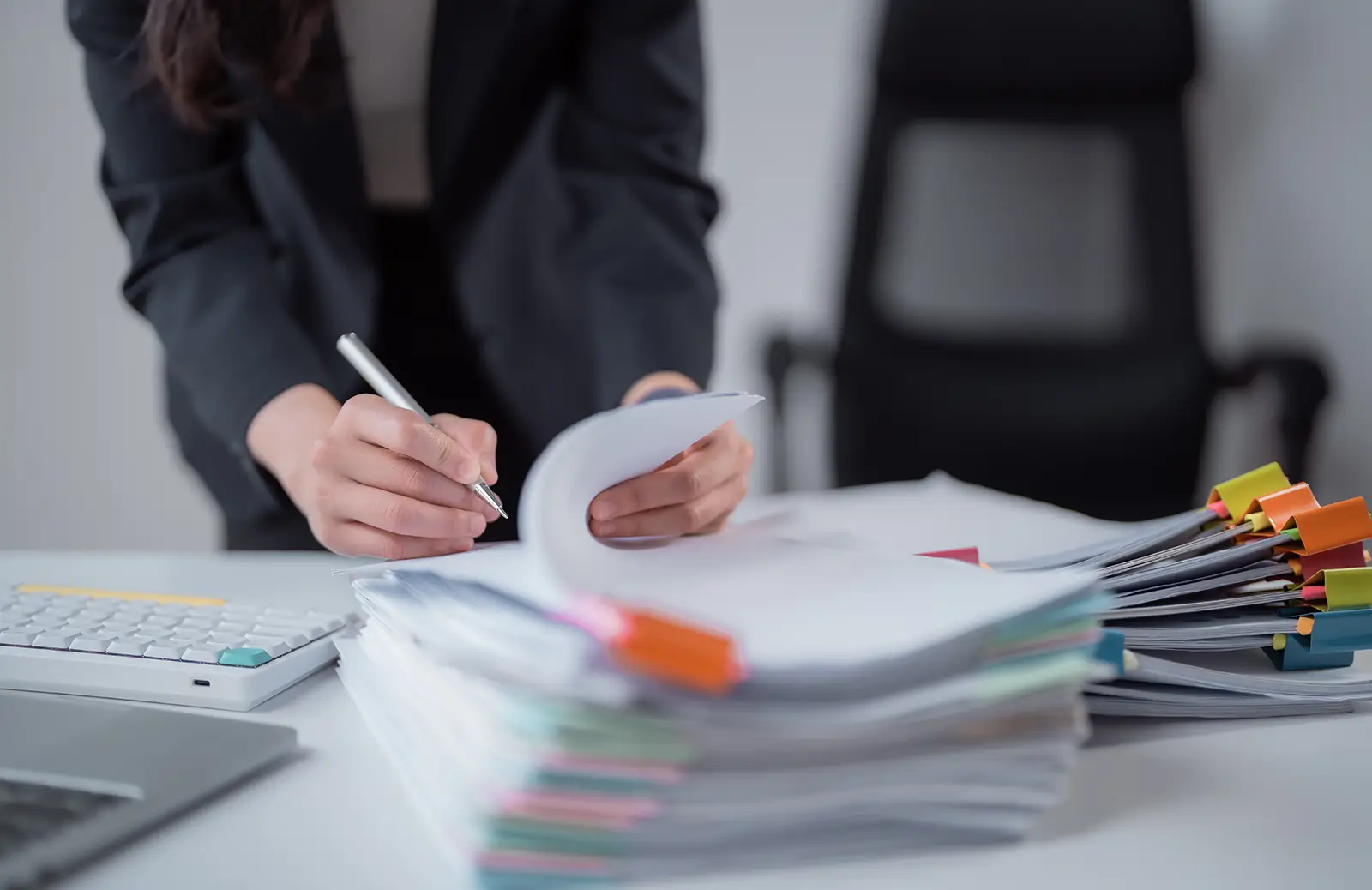 Young professional woman writing notes on stack of files next to a laptop