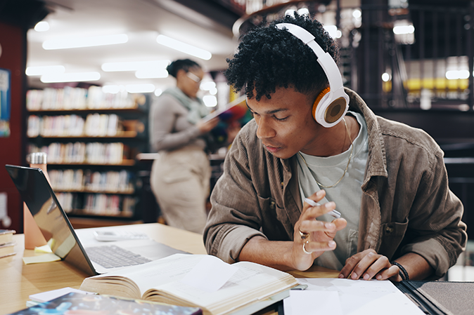 Young man wearing headphones studying with books and a laptop in a library