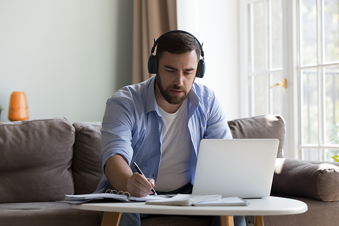 Young man wearing headphones while taking notes in front of his laptop at home