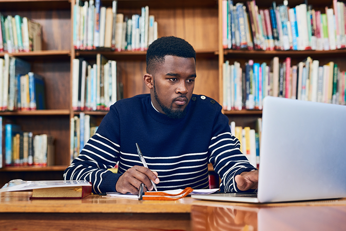 Young man sitting in a library taking notes while working on a laptop