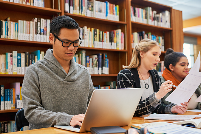 Young man typing on a laptop while in a library with students working at a table nearby