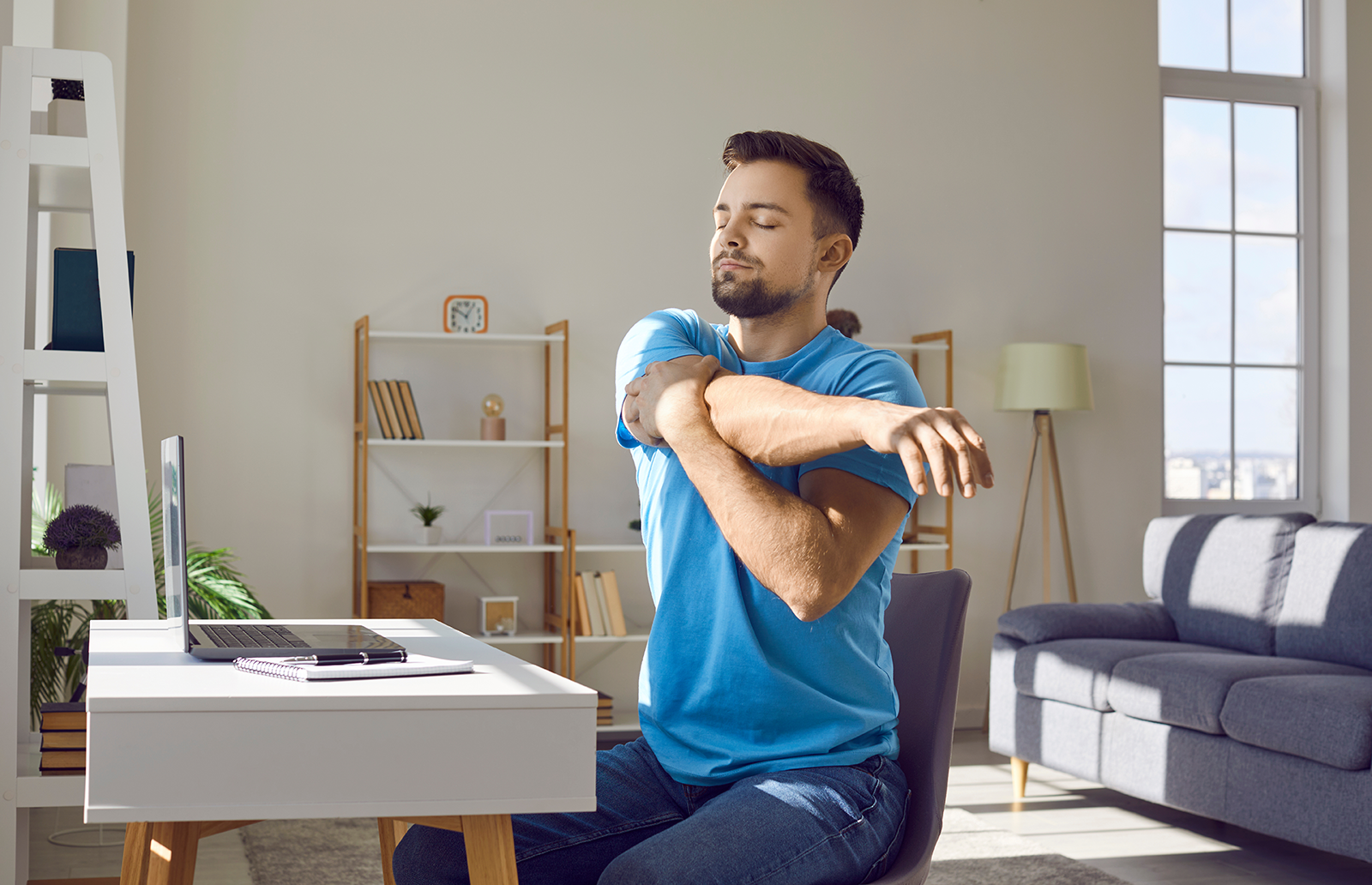 Young man stretching his arms sitting in front of a desk with laptop