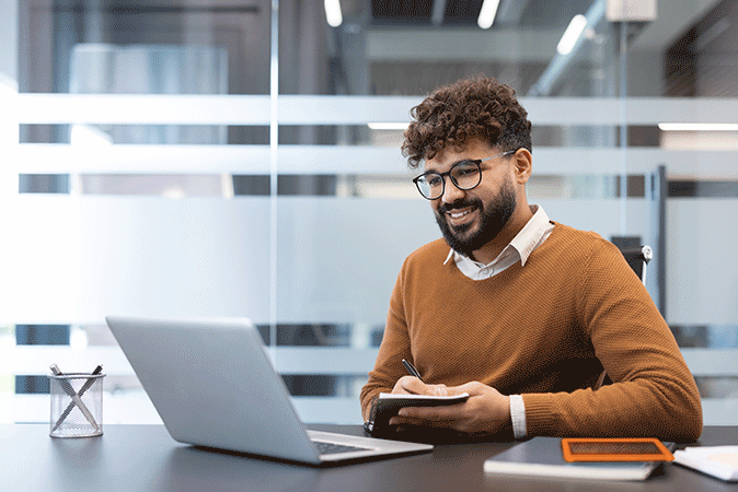 Young man smiling while writing in a notebook looking at his laptop
