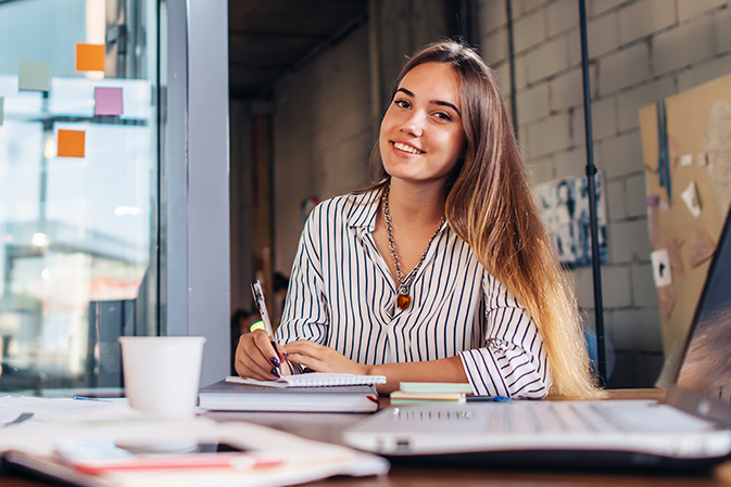 Young confident woman taking notes at her desk in front of a laptop