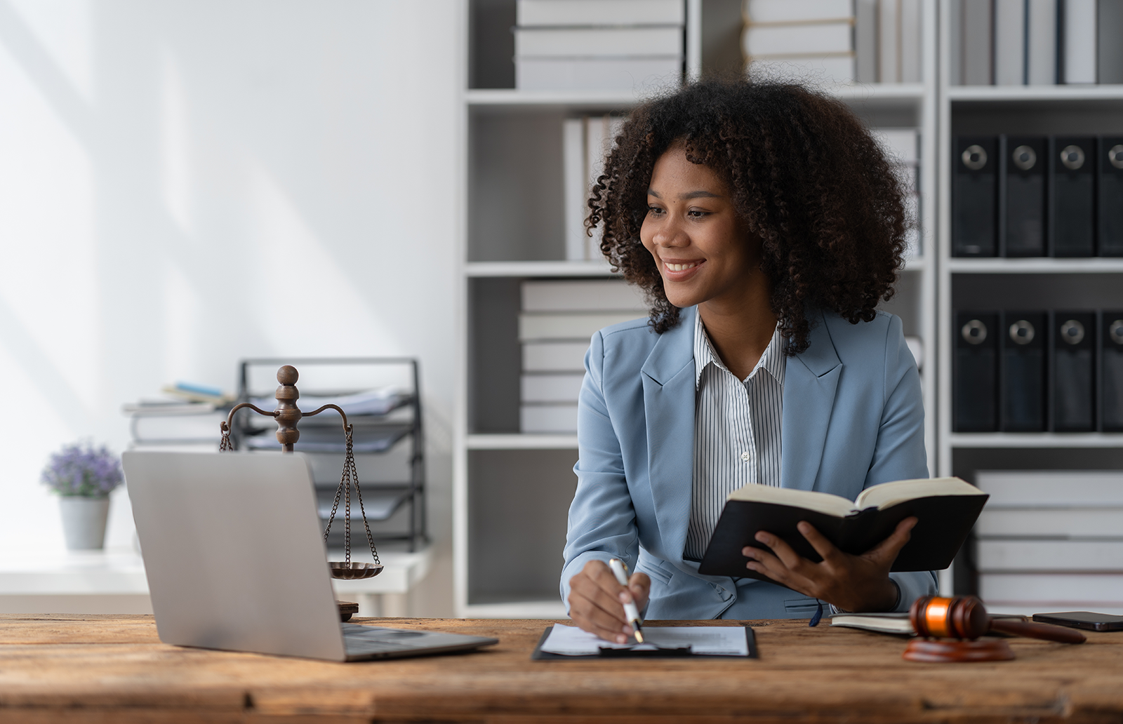 Young woman professional holding a book, taking notes and watching a video on a laptop with books behind her and a judge's gavel on the table