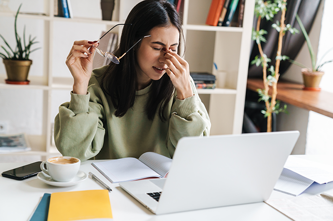 Young woman sitting at a desk in front of a laptop holding the bridge of her nose with a headache