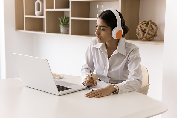 Young woman at home wearing headphones watching video on laptop and taking notes