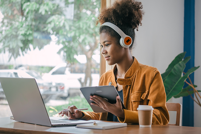 Young woman wearing headphones while holding a tablet and typing on a laptop
