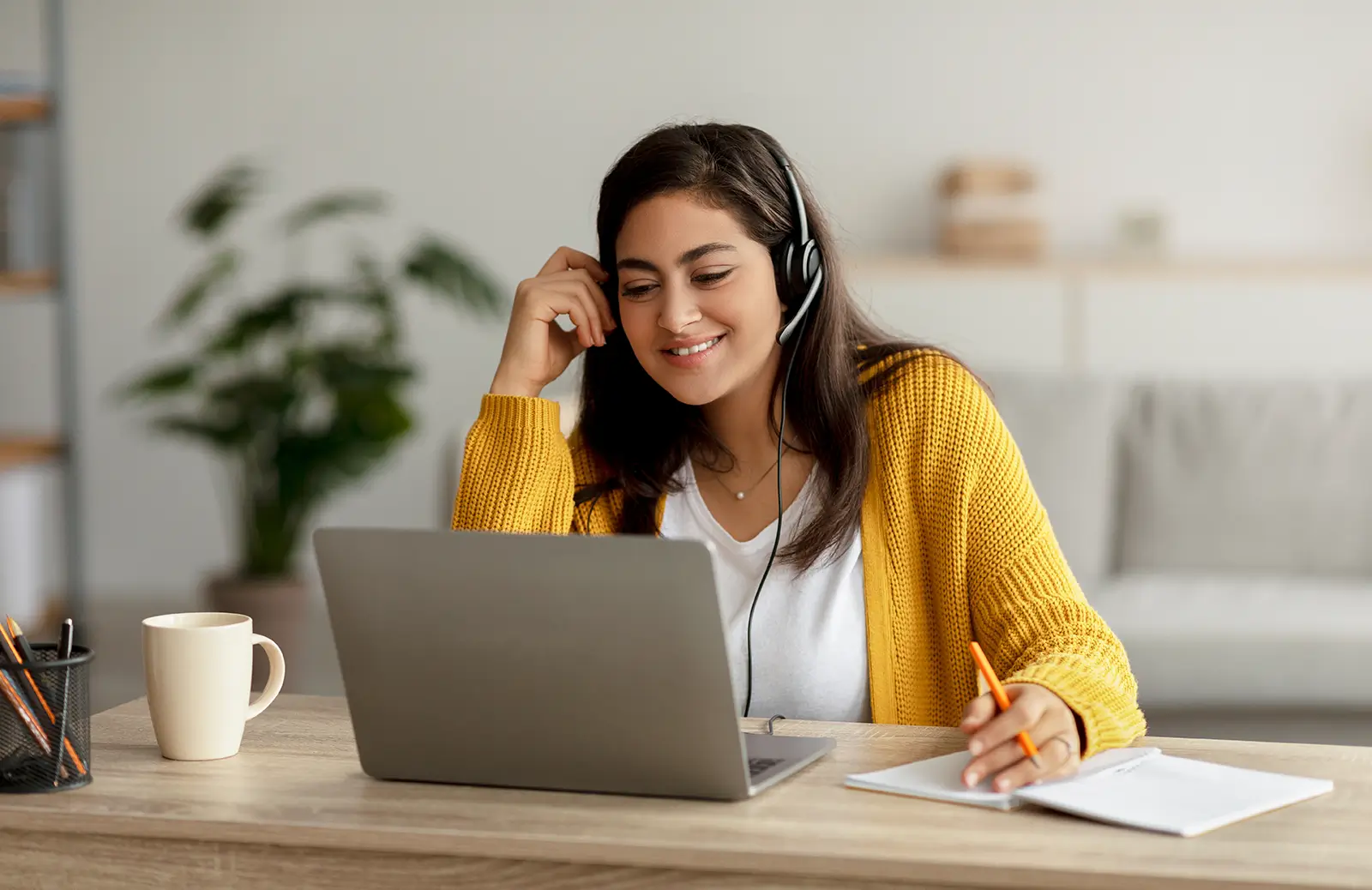 Young woman wearing headphones sitting at a desk watching something on her laptop and taking notes
