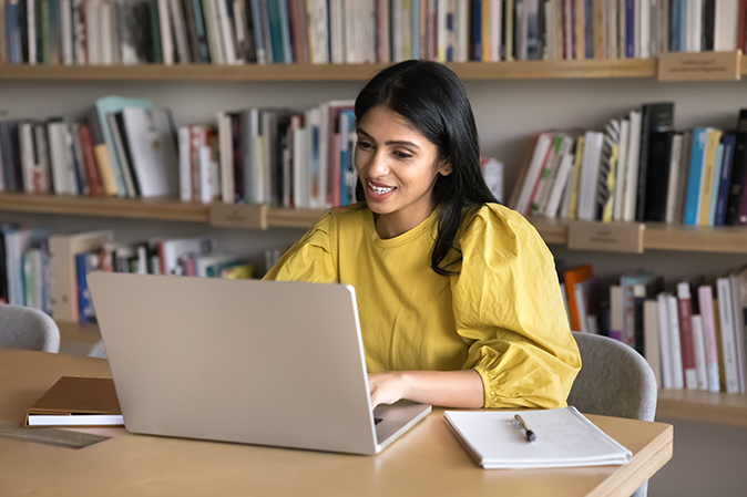 Young woman in a library smiling while typing on a laptop