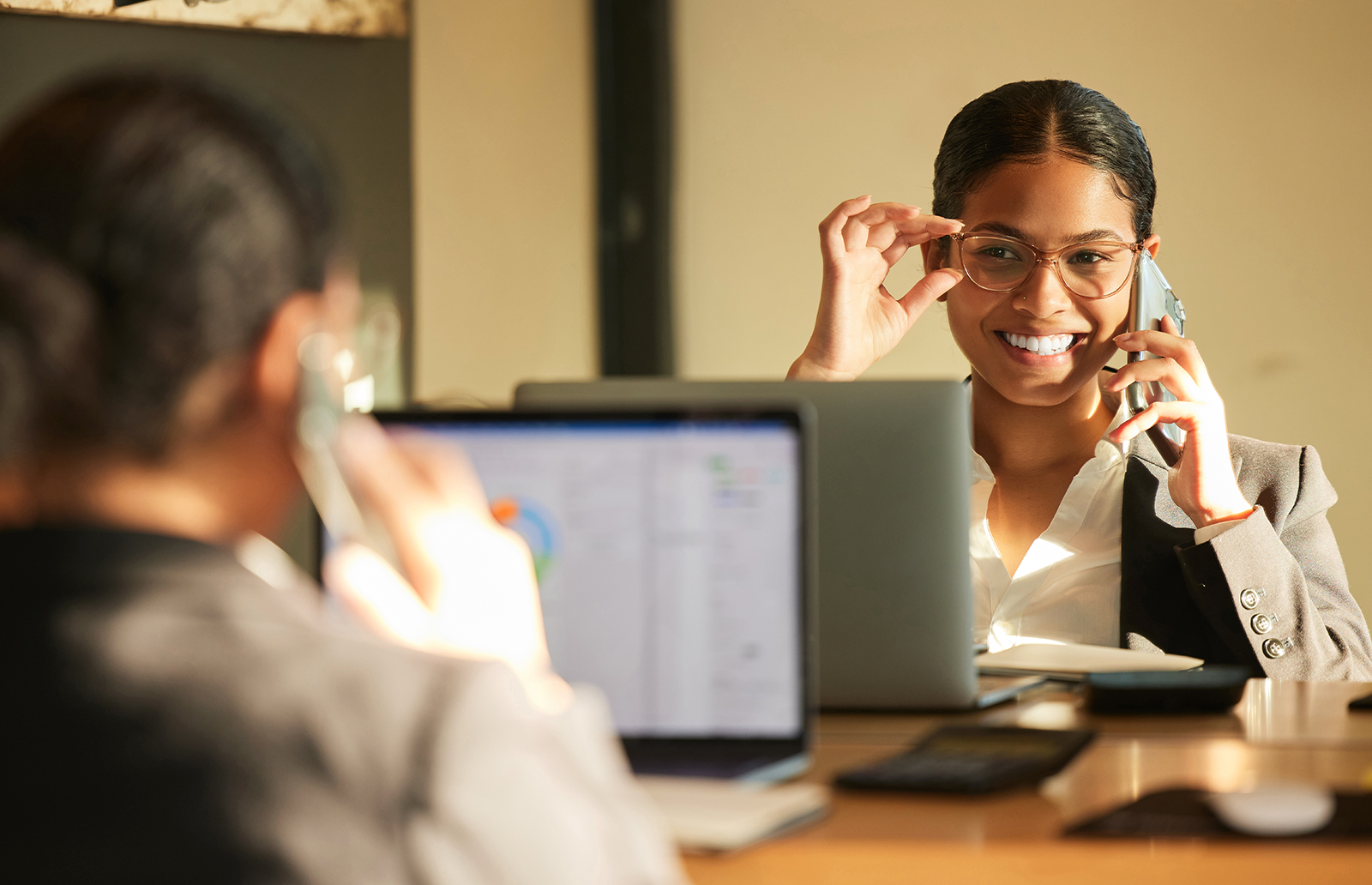 Young woman wearing glasses smiling in the mirror holding a mobile device in front of a laptop