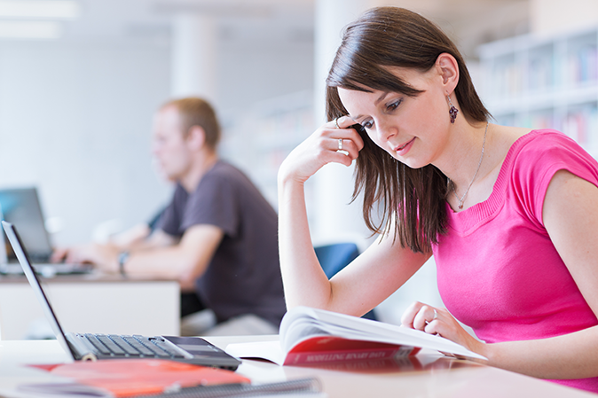 Young woman looking at a book while sitting in class in front of a laptop