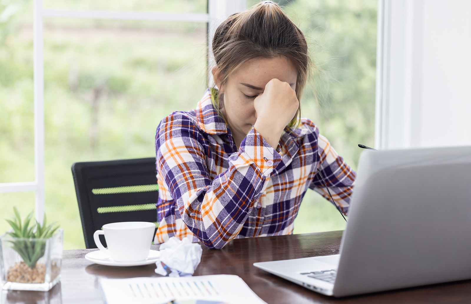 Young woman sitting at a desk in front of a laptop holding the bridge of her nose in frustration