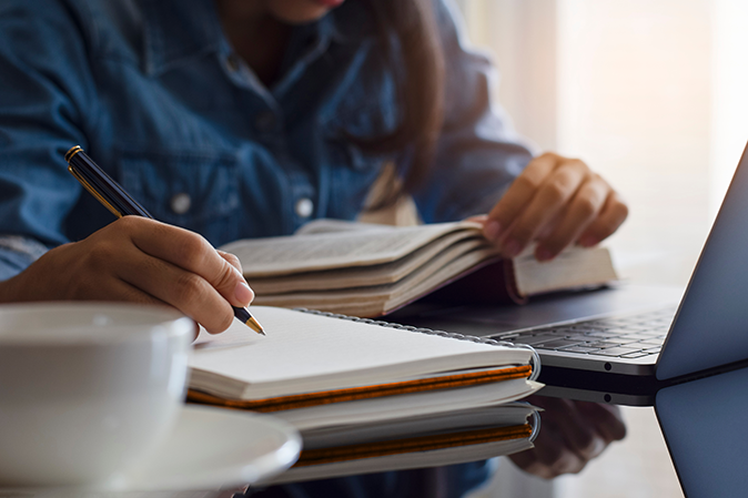 Young woman writing notes while using a textbook in front of a laptop