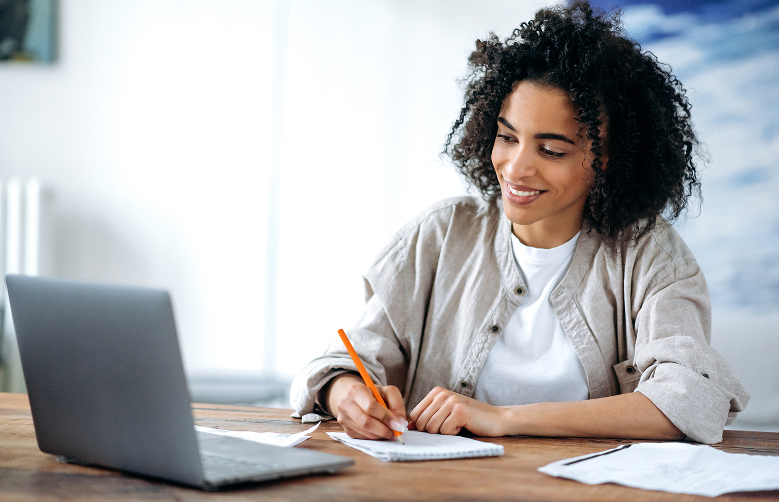 Young woman smiling while writing notes while watching something on a laptop