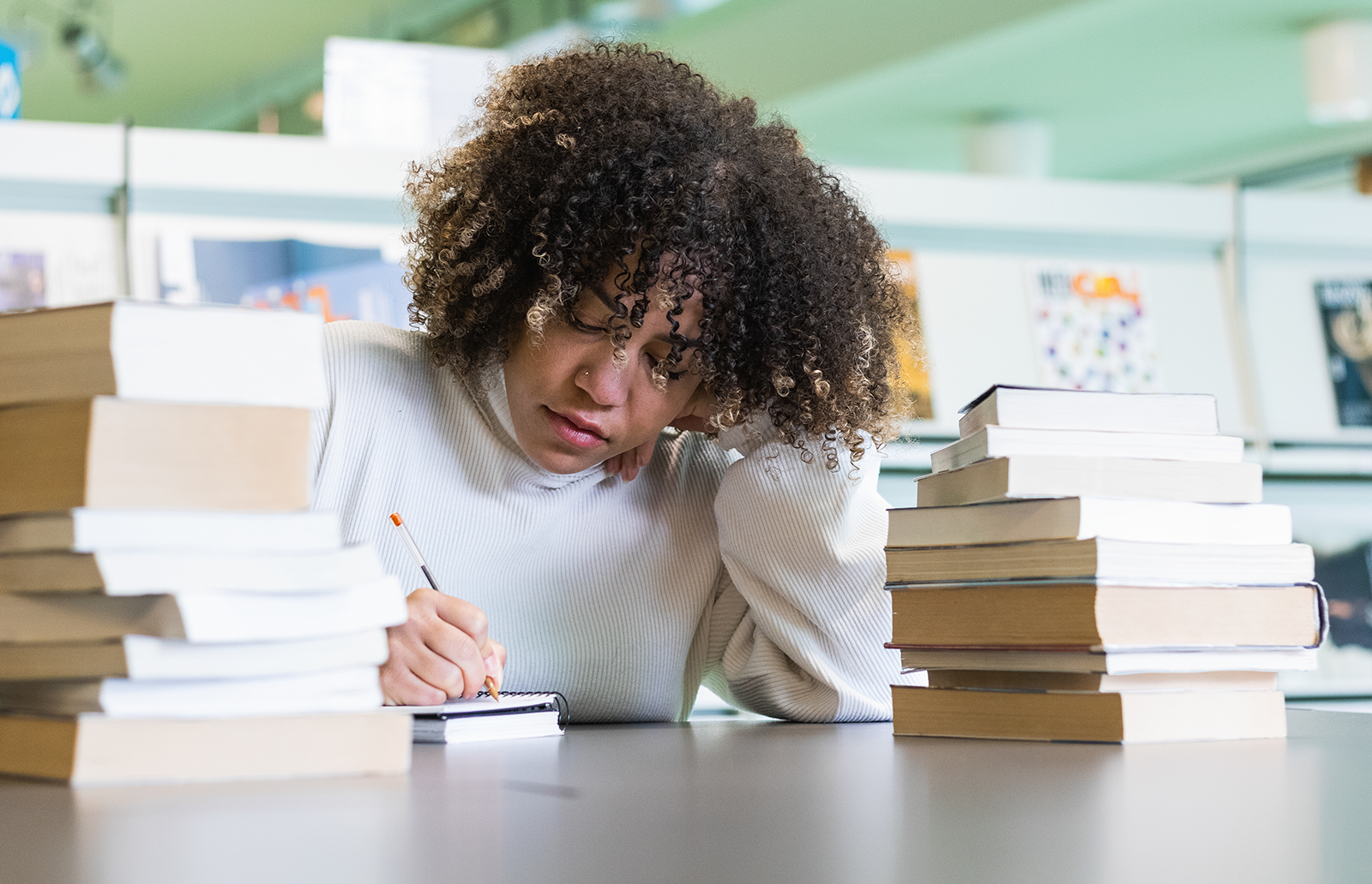 Young woman writing notes in a classroom amongst a couple stacks of books