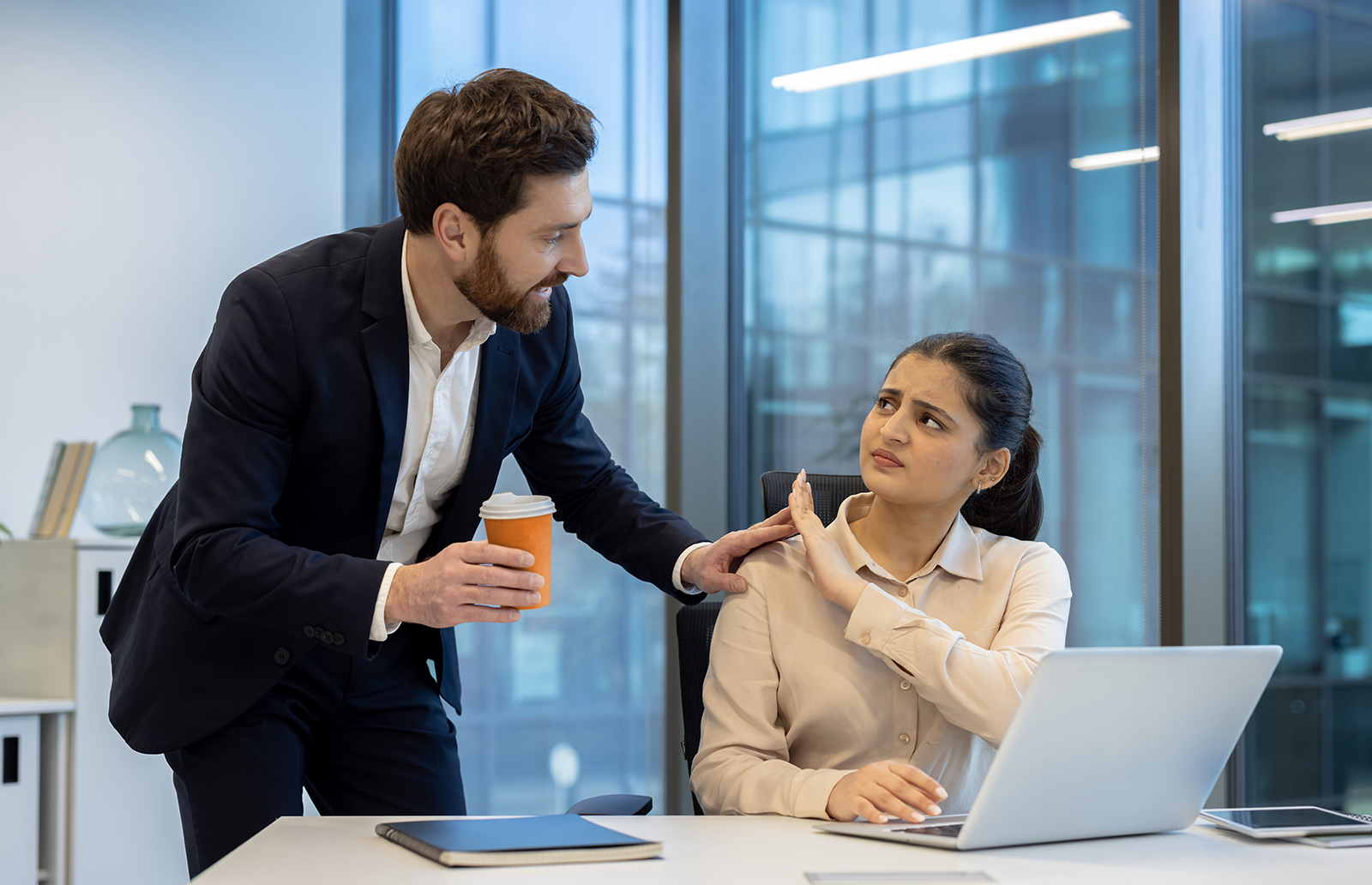 An office setting with a young man putting his hand on the shoulder of a young woman who is trying to push him away