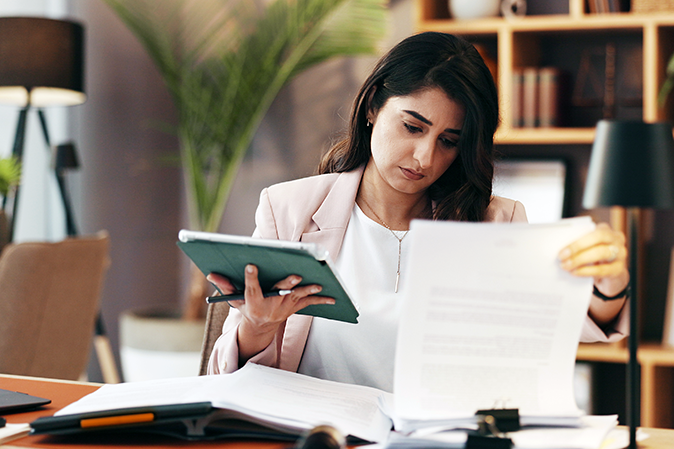 Professional woman holding a tablet while going over paperwork in an office
