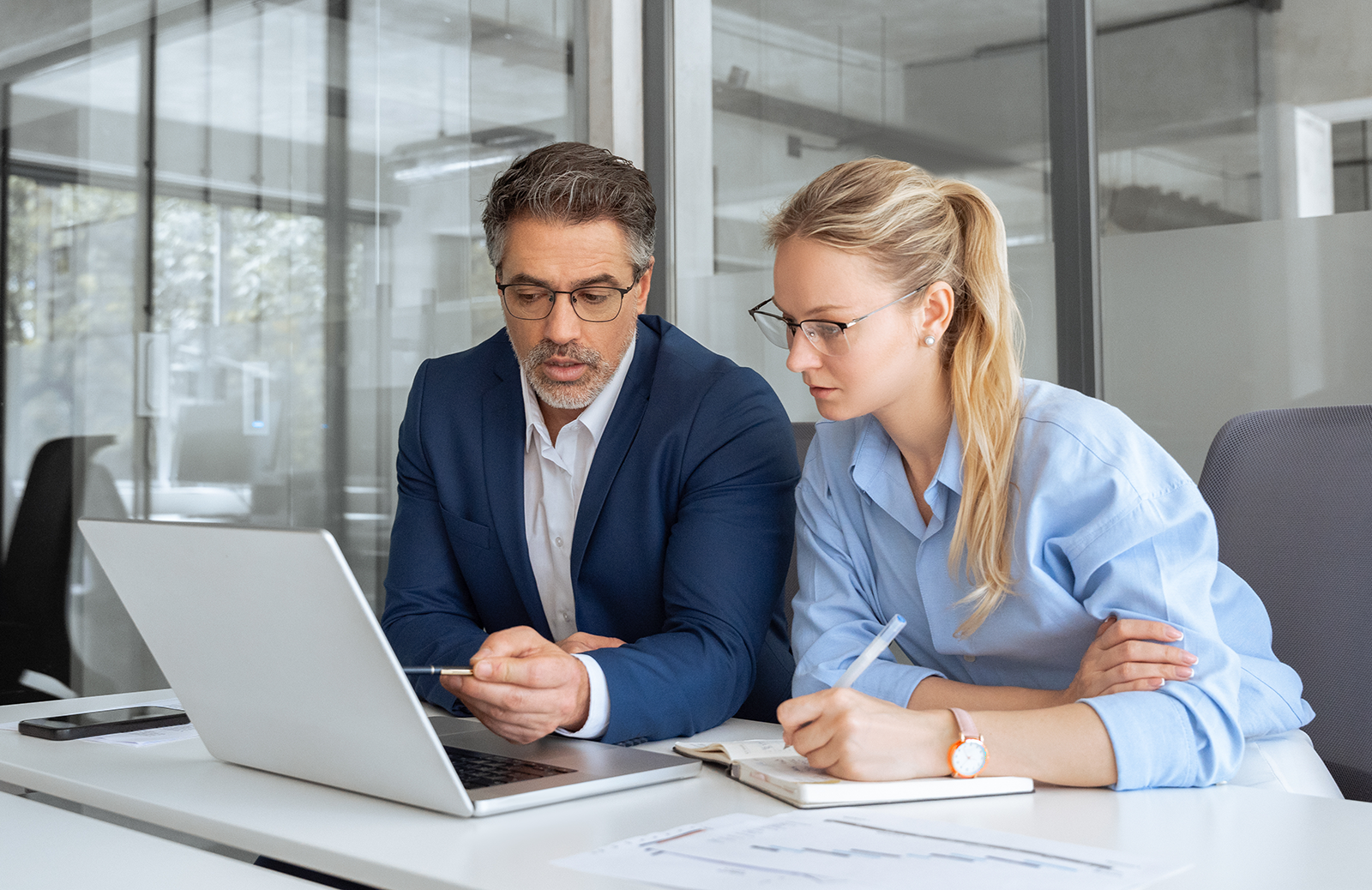 Senior male professional pointing to the screen on a laptop while working with a younger woman is taking notes