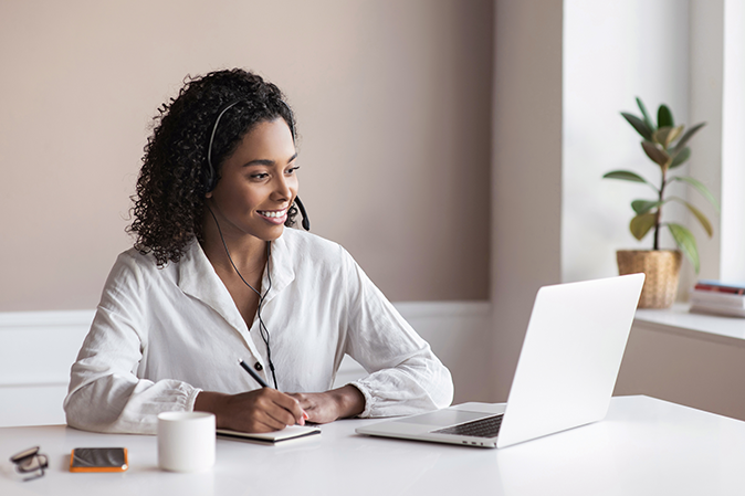 Young woman wearing headphones taking notes in front of a laptop with a coffee, mobile device and glasses nearby