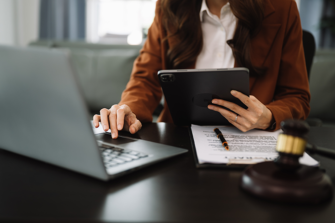 Professional woman sitting at a desk in an office holding a tablet and typing on a laptop