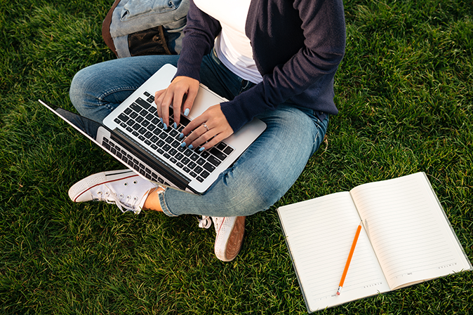 Young woman working on her laptop with open notebook sitting on the grass