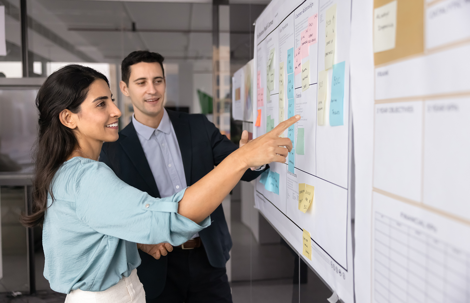 Young woman and young man looking at big whiteboard in an office with multiple projects on it.