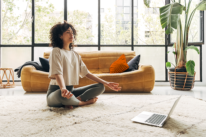 Young woman sitting on floor in living room in a yoga pose in front of laptop