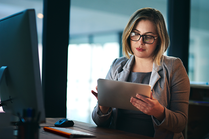 Young woman holding a tablet while sitting at a desk in front of a computer