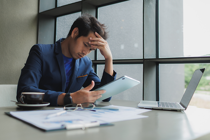 Young professional man with his head in his hand while holding a tablet sitting in front of a laptop