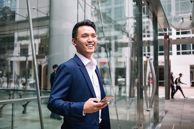 Young man smiling outside an office building holding a mobile device