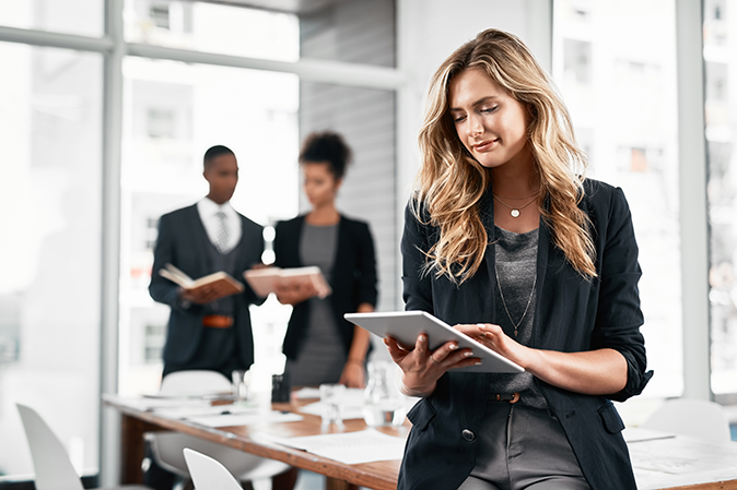 Young professional woman holding a tablet leaning on a table in an office with two co-workers standing behind her