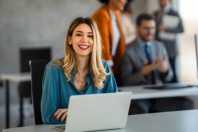 Young professional woman sitting in front of a laptop smiling in an office with coworkers
