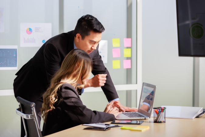 A man and woman working together at a desk, looking at a laptop in a modern office.